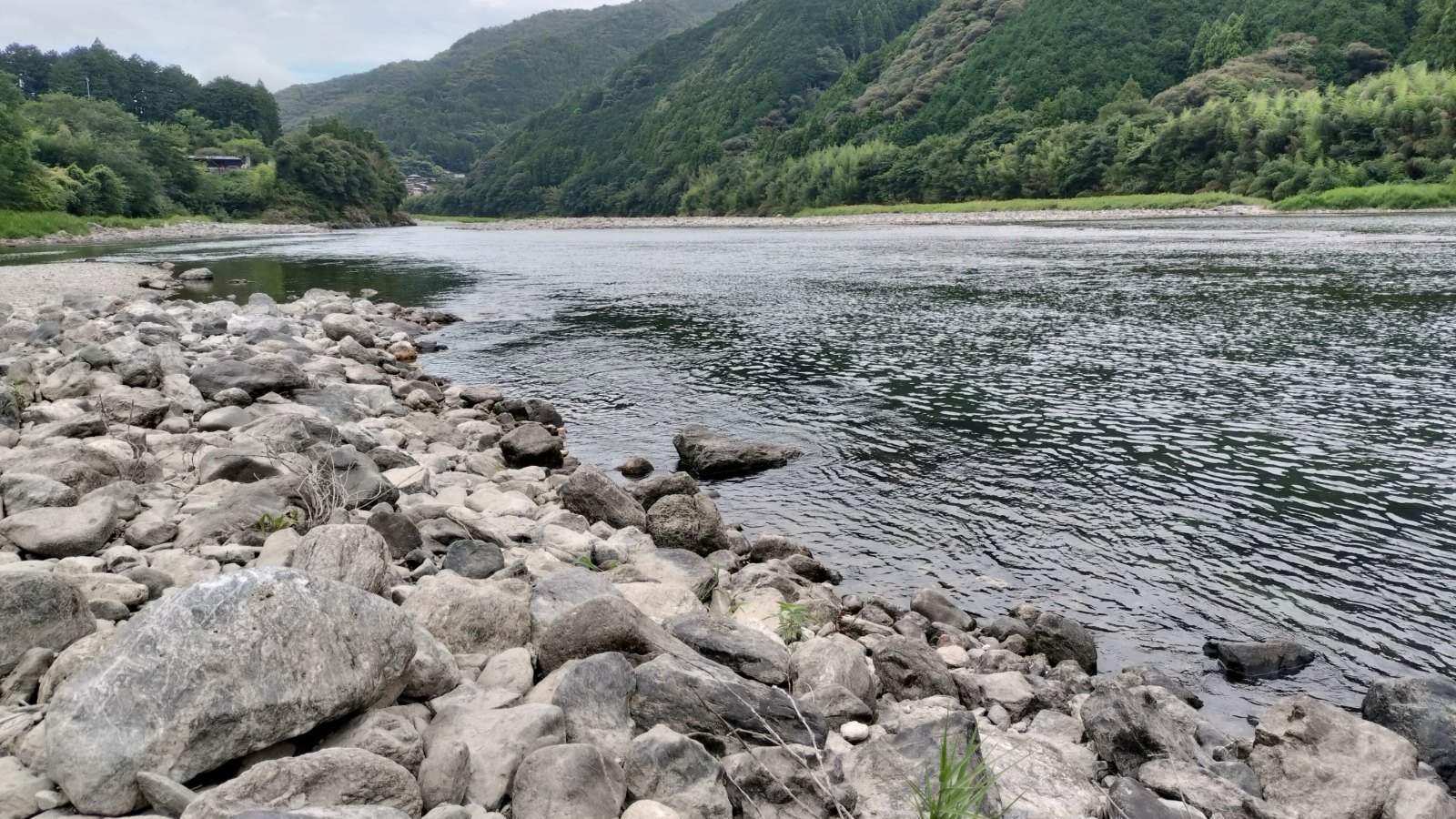 石の河原から見た広い川と、両岸に迫る緑の山。空は曇っている