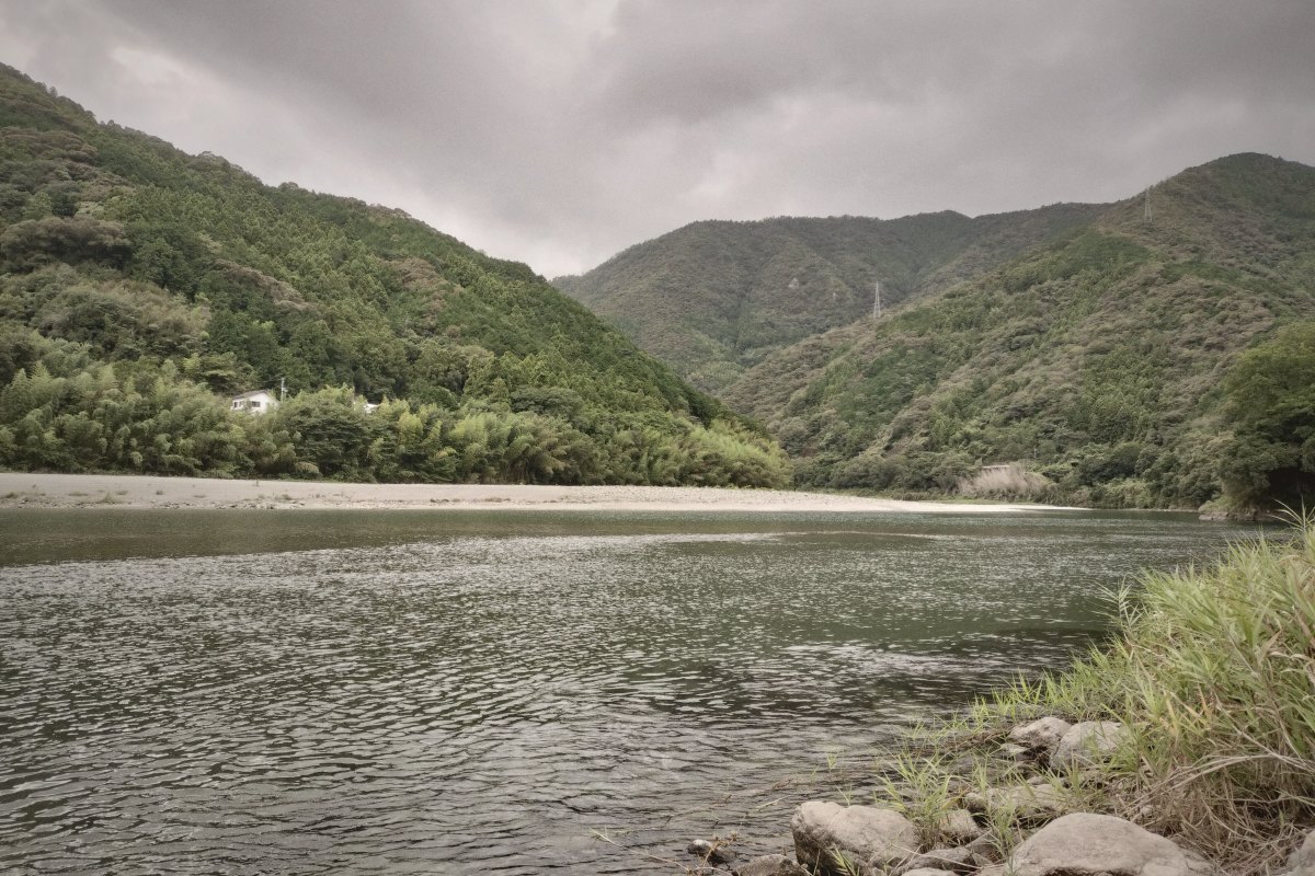 厚い雲の下を流れる広い川と緑の山。対岸には白い河原が続いている