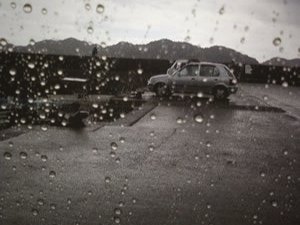 雨粒の付いた車の窓越しに見える、雨に濡れた駐車場と曇り空